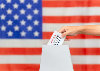 A hand places a vote into a ballot box, set against the backdrop of the United States flag, symbolizing the heart of democracy and inspiring thoughtful US elections analysis. | FintechZoom