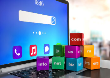 A laptop screen displays a search bar, time 18:16, and app icons. Colorful cubes with domain extensions like ".com" and ".org," hinting at a savvy domain name strategy, are stacked on the keyboard against a blurred office backdrop. | FintechZoom
