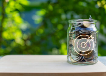 A glass jar filled with coins labeled "Savings" sits on a wooden surface against a blurred green background. A graphic overlay of a target and the word "GOAL" is visible on the jar, emphasizing the journey towards financial objectives. | FintechZoom
