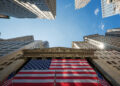 Upward view of Wall Street buildings towering against a clear blue sky, with the facade of the New York Stock Exchange prominently displaying a large American flag. Sunlight reflects off the glass windows of the surrounding skyscrapers, capturing the essence of FintechZoom.com Nasdaq coverage. | FintechZoom