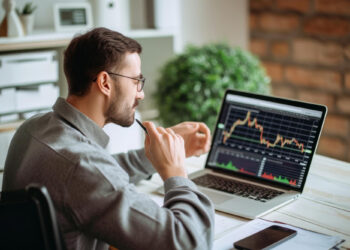A man in a gray shirt and glasses sits at a desk analyzing a fluctuating graph on his laptop, contemplating its implications for cyber security in fintech. A pen held to his chin, he’s surrounded by a smartphone, notebook, and green plant, deep in thought about the future of financial technology. | FintechZoom