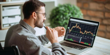 A man in a gray shirt and glasses sits at a desk analyzing a fluctuating graph on his laptop, contemplating its implications for cyber security in fintech. A pen held to his chin, he’s surrounded by a smartphone, notebook, and green plant, deep in thought about the future of financial technology. | FintechZoom