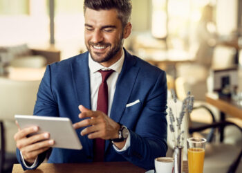 A man in a blue suit and red tie sits at a table in a café, smiling while using a tablet loaded with collaboration tools. A cup of coffee and a glass of orange juice sit beside him. The setting is bright with natural light filtering through the windows. | FintechZoom