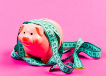 A pink piggy bank wrapped in a blue measuring tape on a pink background, symbolizing how non-profit organizations manage savings and budgeting effectively, even without big budgets. | FintechZoom