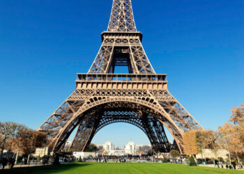 The Eiffel Tower stands tall against a clear blue sky, surrounded by lush green grass and autumn-colored trees. As one of the highlights in one of the best European countries to visit, France, this iconic iron structure dominates the landscape, with a few people visible at its base. | FintechZoom