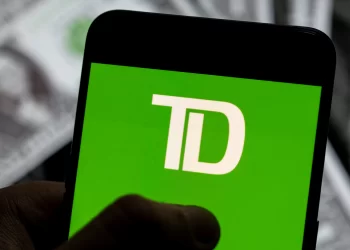A smartphone displays the TD Bank Online logo on a green screen. In the background, there's a blurred image of U.S. dollar bills, emphasizing seamless and secure digital banking. | FintechZoom
