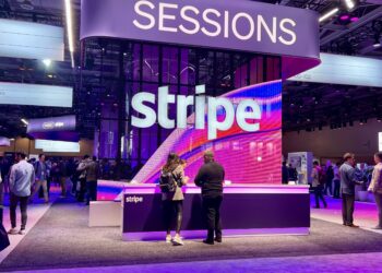 A booth at a conference with a large "Stripe" sign and "Sessions" written above. People are gathered around with a purple and white color scheme throughout the space. | FintechZoom