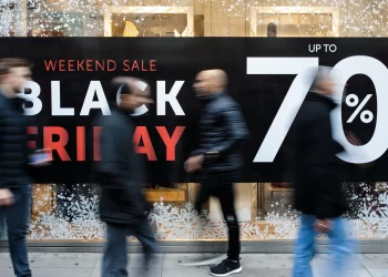 Four people walk past a storefront window displaying a "Weekend Sale Black Friday 2024 Up to 70%" sign. The window is decorated with white snowflake patterns, and the scene is bustling with movement. | FintechZoom