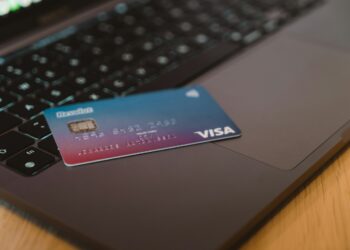 A Visa credit card is placed on the keyboard of an open laptop, suggesting online transactions. The card has a gradient blue and purple design and the contactless payment symbol. The scene is set on a wooden surface. | FintechZoom