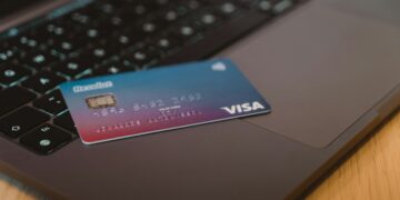 A Visa credit card is placed on the keyboard of an open laptop, suggesting online transactions. The card has a gradient blue and purple design and the contactless payment symbol. The scene is set on a wooden surface. | FintechZoom