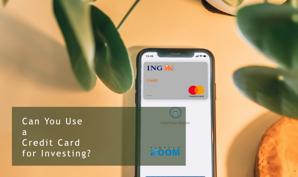 A smartphone displaying credit card options is placed on a desk with green leaves around it. Text on the image reads, "Can You Use a Credit Card for Investing?. | FintechZoom