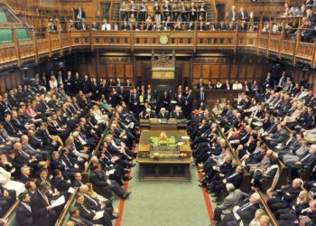 A busy parliamentary chamber buzzes with urgency as politicians discuss the proposed law to cap Oasis ticket costs. The room, with its wooden paneling and large benches, sees officials gathered around a central table. The atmosphere remains serious and formal amidst the heated debate. | FintechZoom