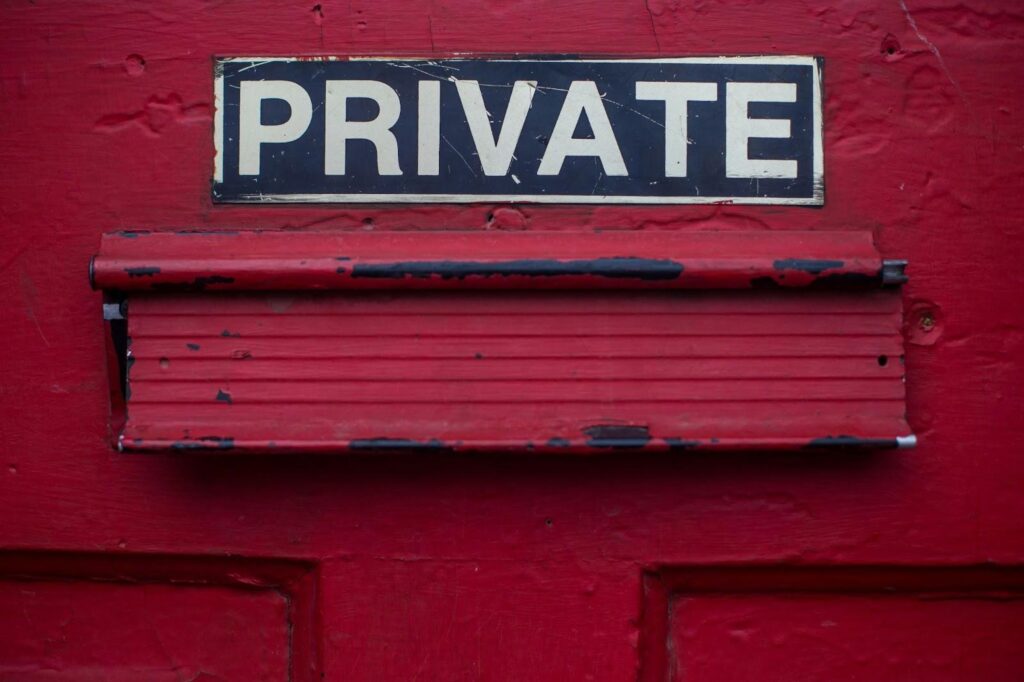 A weathered red door with a mail slot, featuring a sign above it that reads "PRIVATE" in bold white letters on a black background. The door's paint is chipping around the edges, giving it a rustic appearance. | FintechZoom