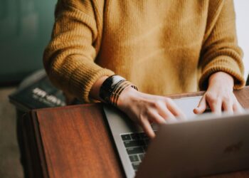 Person in a mustard sweater typing on a laptop at a wooden desk. The focus is on their hands and sweater, with a glimpse of books nearby. The setting appears casual and cozy. | FintechZoom