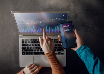 Two people use technology for financial analysis. One points to a laptop screen displaying a colorful graph with "Yesterday" and "Today." The other holds a phone showing stock prices. They are at a table with a dark surface. | FintechZoom