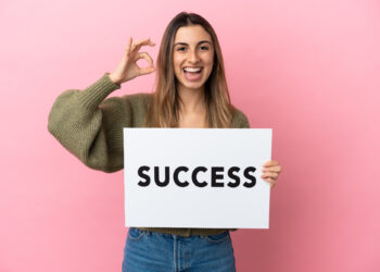A person smiling and holding a sign that reads "SUCCESS" against a pink background. They are making an "OK" hand gesture with their other hand. | FintechZoom