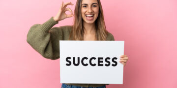 A person smiling and holding a sign that reads "SUCCESS" against a pink background. They are making an "OK" hand gesture with their other hand. | FintechZoom