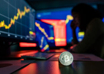 A Bitcoin coin stands in focus on a table surrounded by computer screens displaying financial charts and data. In the dimly lit room, a person sits in the background, appearing pensive as they contemplate betting strategies amidst the fluctuating digital landscapes. | FintechZoom