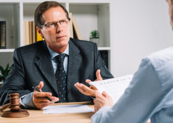A serious-looking man in a suit, likely a lawyer for a personal injury lawsuit, sits at a desk with a gavel, discussing details with another person holding a contract. Shelves filled with books and the presence of a potted plant add depth to the professional setting. | FintechZoom