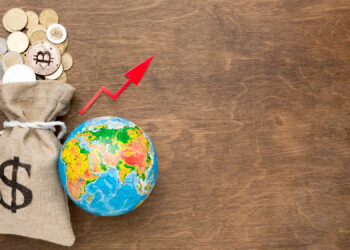 A burlap sack with a dollar sign is filled with various coins, including a Bitcoin, next to a small globe. A red arrow points upward diagonally, indicating growth, on a wooden surface. | FintechZoom