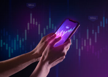 Hands holding a smartphone with a glowing screen showing graphs, against a dark background with financial charts and "SELL" labels, indicating stock market analysis or trading. | FintechZoom