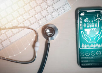 A smartphone displaying ecology-themed graphics with a wind turbine, trees, and globe icons rests beside a keyboard and stethoscope, subtly aligning fintech in health with environmental awareness for a healthier future. | FintechZoom