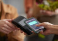 A person uses a smartphone to make a contactless payment on a card reader. Another person holds the card reader steady. Both are indoors, and a blurred background suggests a commercial environment. | FintechZoom
