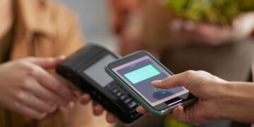 A person uses a smartphone to make a contactless payment on a card reader. Another person holds the card reader steady. Both are indoors, and a blurred background suggests a commercial environment. | FintechZoom