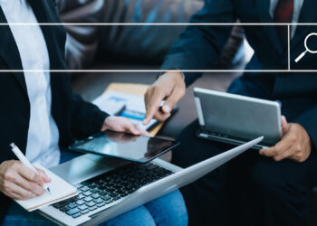 Two people sitting on a sofa are working with digital tablets and a laptop, with a notebook for notes. A search bar icon hovers above them, indicating online research or browsing. | FintechZoom