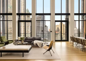 A modern, spacious living room with large windows showcasing a cityscape. It has a minimalist design with green and gray sofas, a wooden chair, coffee table, and a long dining table with chairs. Natural light floods the space. | FintechZoom