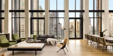 A modern, spacious living room with large windows showcasing a cityscape. It has a minimalist design with green and gray sofas, a wooden chair, coffee table, and a long dining table with chairs. Natural light floods the space. | FintechZoom