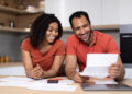 A smiling couple reviews financial documents at a kitchen table. They are holding papers and wearing matching orange shirts. A tablet and a calculator are on the table. The background shows kitchen shelves with various items. | FintechZoom