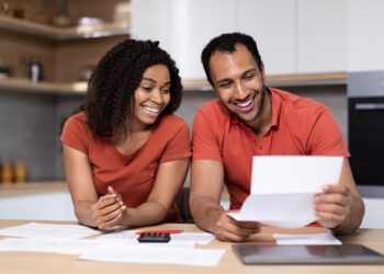 A smiling couple reviews financial documents at a kitchen table. They are holding papers and wearing matching orange shirts. A tablet and a calculator are on the table. The background shows kitchen shelves with various items. | FintechZoom