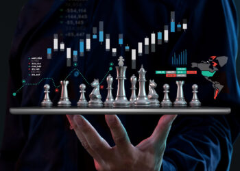 A person holds a tablet showing a chessboard with silver pieces, overlaid with stock market graphs and digital data from FintechZoom.com. The dark background emphasizes the strategic link between chess and financial markets. | FintechZoom