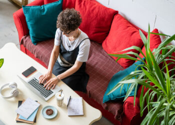 A person with curly hair is working on a laptop while sitting on a red couch. There's a notebook, a pen, a phone, headphones, and a glass of coffee with a cookie on the white table. Green plants are nearby. | FintechZoom