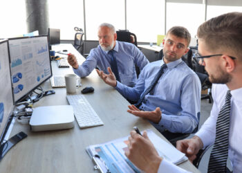 Three men in business attire are sitting at a desk with multiple computer screens displaying charts and graphs. They appear to be in a discussion, with papers and a coffee cup on the table in a bright office setting. | FintechZoom