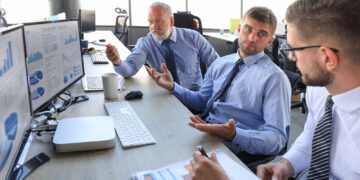 Three men in business attire are sitting at a desk with multiple computer screens displaying charts and graphs. They appear to be in a discussion, with papers and a coffee cup on the table in a bright office setting. | FintechZoom