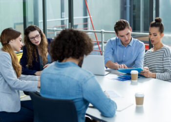 A group of people sitting around a table in a modern office setting. They are engaged in a discussion, with papers, a laptop, and coffee cups visible on the table. Large windows are in the background, allowing natural light to enter. | FintechZoom