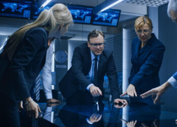 A group of people in business attire stand around a reflective table in a dimly lit conference room, intently pointing at various spots on the table. Multiple screens and clocks are visible in the background. | FintechZoom