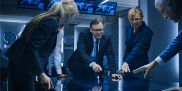 A group of people in business attire stand around a reflective table in a dimly lit conference room, intently pointing at various spots on the table. Multiple screens and clocks are visible in the background. | FintechZoom