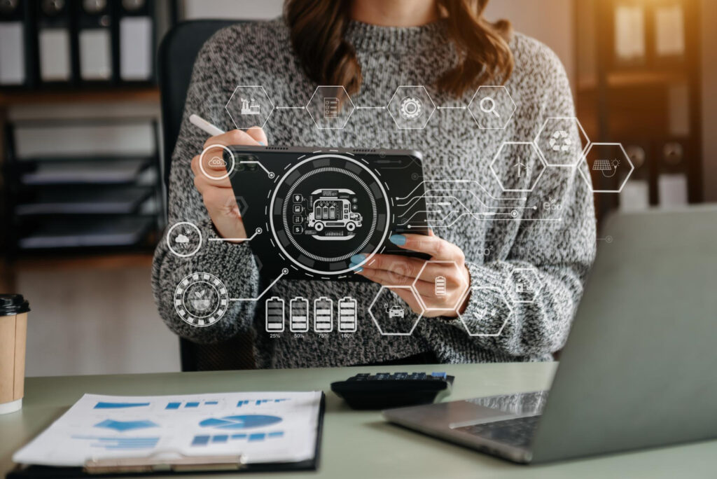 A person in a gray sweater uses a tablet at a desk. Transparent digital icons and hexagons overlay the image, symbolizing technology and automation in finance. Nearby are documents with graphs, a calculator, and a takeaway coffee cup, highlighting the seamless integration of data analysis. | FintechZoom