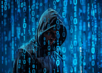 A person in a hooded jacket stands against a digital background of blue binary code. The numbers 1 and 0 are prominently displayed, evoking themes of hacking or cyber technology. The image conveys a mysterious and technological atmosphere. | FintechZoom