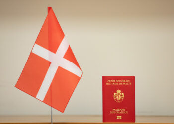 A small red flag with a white cross stands next to a red diplomatic passport with the inscription "Ordre Souverain Militaire de Malte" on the cover. The background is a plain off-white wall. | FintechZoom