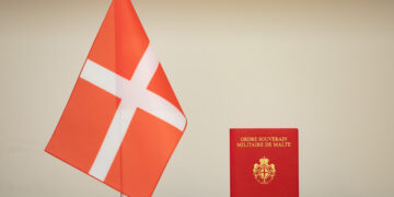 A small red flag with a white cross stands next to a red diplomatic passport with the inscription "Ordre Souverain Militaire de Malte" on the cover. The background is a plain off-white wall. | FintechZoom