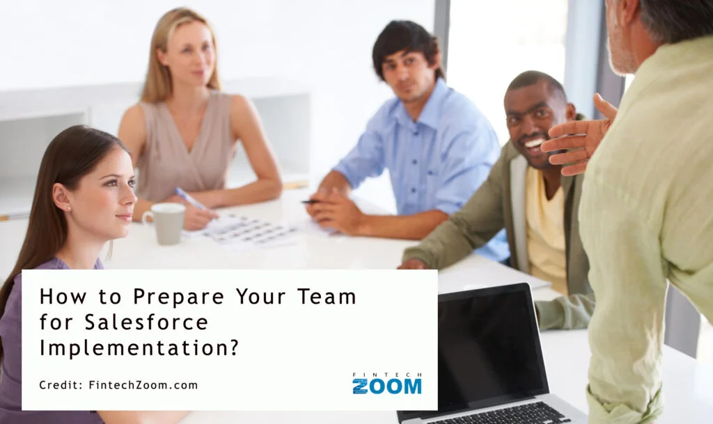 A diverse group of four people sits around a table in a meeting, attentively listening to a standing presenter. The text on the image reads, "How to Prepare Your Team for Seamless Salesforce Implementation?" Credit: FintechZoom.com. | FintechZoom