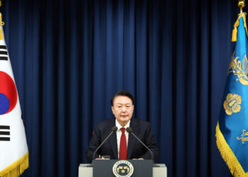 President Yoon Suk Yeol of South Korea stands at a podium with microphones,Declares Martial Law. He is flanked by two flags: the flag of South Korea on the left and another flag with a blue background and gold emblem on the right. A dark curtain serves as the backdrop. | FintechZoom