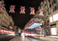 A nighttime street scene features a red double-decker bus creating light trails as it drives past. Union Jack flags adorn the road, and lit-up buildings surround pedestrians strolling along the sidewalk, their conversations occasionally touching on investing in the FTSE 100 as indicated by FintechZoom.com. | FintechZoom