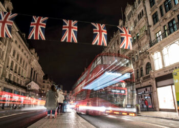 A nighttime street scene features a red double-decker bus creating light trails as it drives past. Union Jack flags adorn the road, and lit-up buildings surround pedestrians strolling along the sidewalk, their conversations occasionally touching on investing in the FTSE 100 as indicated by FintechZoom.com. | FintechZoom