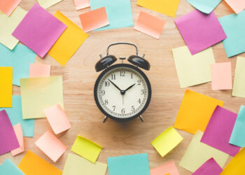An analog alarm clock sits on a wooden surface, surrounded by colorful sticky notes—pink, yellow, orange, blue, and green. The clock shows 10:10. This vibrant display symbolizes effective time management in remote work, each note a reminder to stay organized and productive. | FintechZoom