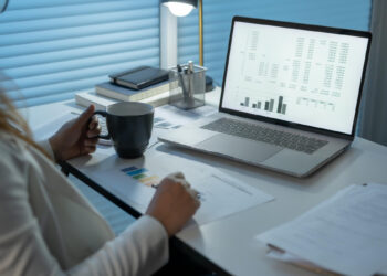 A person holding a coffee mug sits at a desk with a laptop displaying data charts related to expense management. Papers, books, and a pen are scattered on the desk. A desk lamp illuminates the workspace, creating an atmosphere that is calm and focused on work or study. | FintechZoom